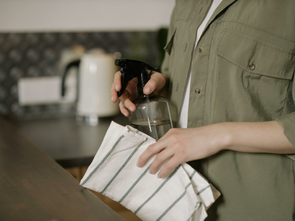Un geste simple pour un intérieur toujours frais Personne nettoyant un plan de travail avec un spray naturel et un torchon réutilisable, dans une cuisine à l'ambiance douce et épurée.